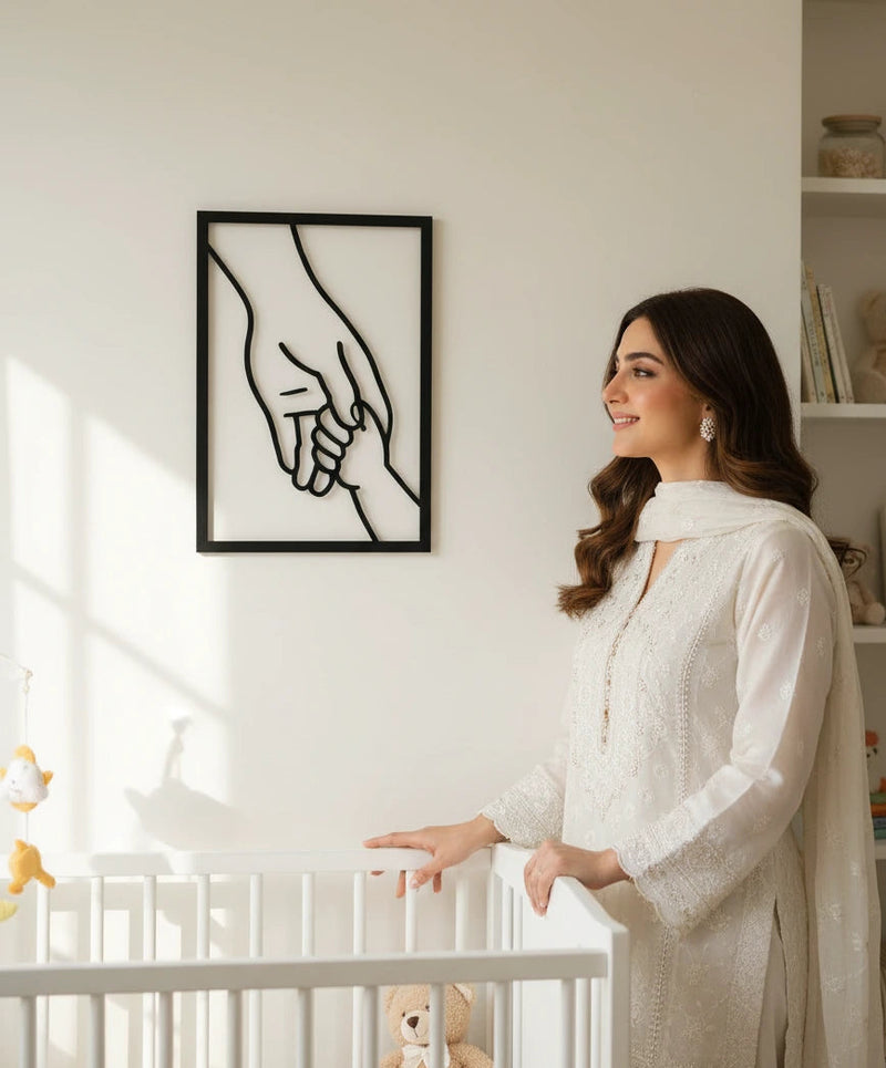 Woman standing in a nursery room with a crib and teddy bear, looking at a framed picture of a mother's hand holding a baby's hand.